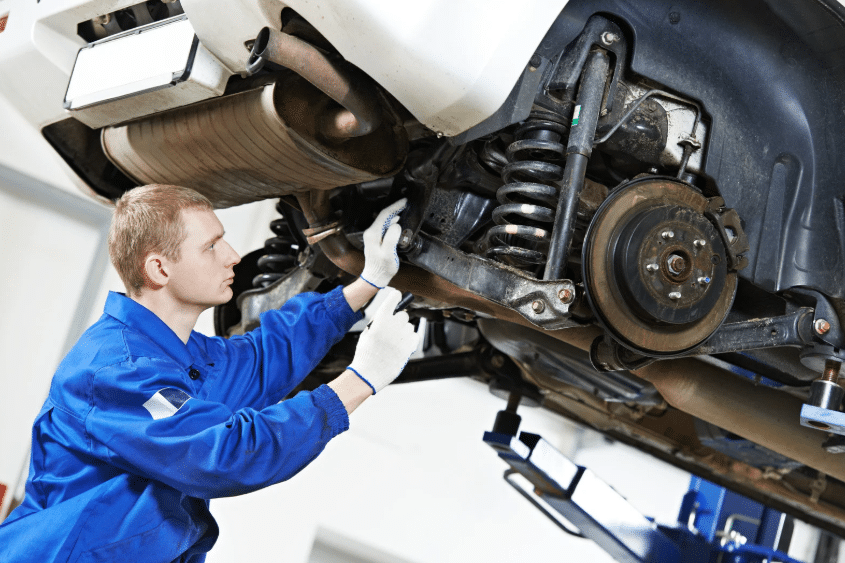 Auto technician performing a suspension check on a lifted vehicle in a repair shop.